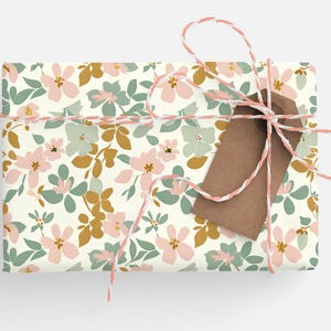 Op de afbeelding: Een cadeau verpakt in wit bloemmotief verpakkingspapier met een bruin cadeaukaartje vastgebonden met touw. Het cadeaupapier heeft een roze, groene en gouden bloemenpatroon.