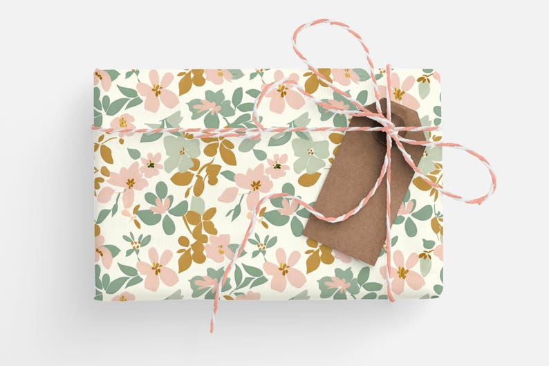 Op de afbeelding: Een cadeau verpakt in wit bloemmotief verpakkingspapier met een bruin cadeaukaartje vastgebonden met touw. Het cadeaupapier heeft een roze, groene en gouden bloemenpatroon.