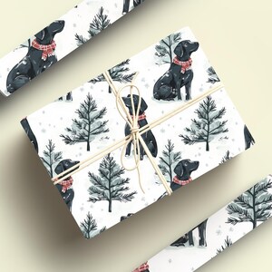 K&ouml;nnte beinhalten: Wei&szlig;es Geschenkpapier mit einem sich wiederholenden Muster aus schwarzen Labradoren mit roten Schottenkaro-Schals und stilisierten Tannen. Das verpackte Geschenk ist mit Bindfaden gebunden, und zwei Rollen passenden Papiers sind ebenfalls sichtbar.