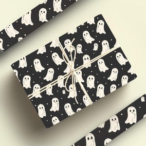 May include: A black gift box with a white ghost pattern wrapped with twine. The box is surrounded by rolls of wrapping paper with the same ghost pattern.