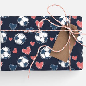 K&ouml;nnte beinhalten: Eine dunkelblaue Geschenkpapierrolle mit einem sich wiederholenden Muster aus wei&szlig;en Fu&szlig;b&auml;llen und rosafarbenen Herzen. Das Geschenk ist mit einem wei&szlig;en und roten Band und einem braunen Geschenkanh&auml;nger gebunden.