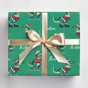 May include: Green wrapping paper with a repeating pattern of dinosaurs wearing Santa hats and riding skateboards. The paper is tied with a gold ribbon and the name "Hudson" is printed on the paper.