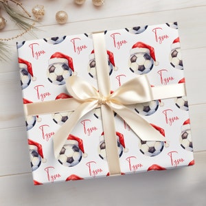 May include: White gift wrap with a repeating pattern of soccer balls wearing Santa hats. The name "Tyson" is repeated throughout the pattern in red.