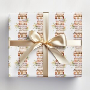 May include: White gift wrap with a repeating pattern of stacks of books and floral sprigs. A gold ribbon is tied around the box with a bow.