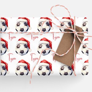 May include: White wrapping paper with a repeating pattern of soccer balls wearing Santa hats. The name "Tyson" is printed in red on the paper. A brown gift tag is tied to the paper with white and pink twine.