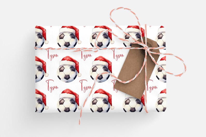 May include: White wrapping paper with a repeating pattern of soccer balls wearing Santa hats. The name "Tyson" is printed in red on the paper. A brown gift tag is tied to the paper with white and pink twine.
