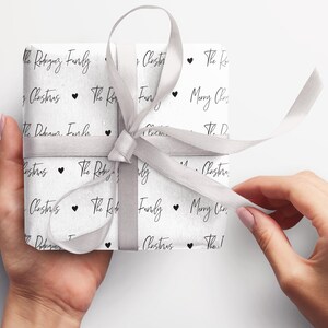 May include: A white gift box wrapped in white paper with a black and white pattern. The pattern includes the text "The Rodriguez Family" and "Merry Christmas" repeated throughout. A silver ribbon is tied around the box.