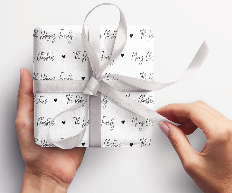 May include: A white gift box wrapped in white paper with a black and white pattern. The pattern includes the text "The Rodriguez Family" and "Merry Christmas" repeated throughout. A silver ribbon is tied around the box.