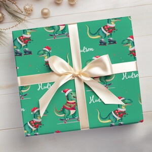 May include: Green wrapping paper with a repeating pattern of dinosaurs wearing Santa hats and riding skateboards. The paper is tied with a gold ribbon and the name "Hudson" is written on the paper.