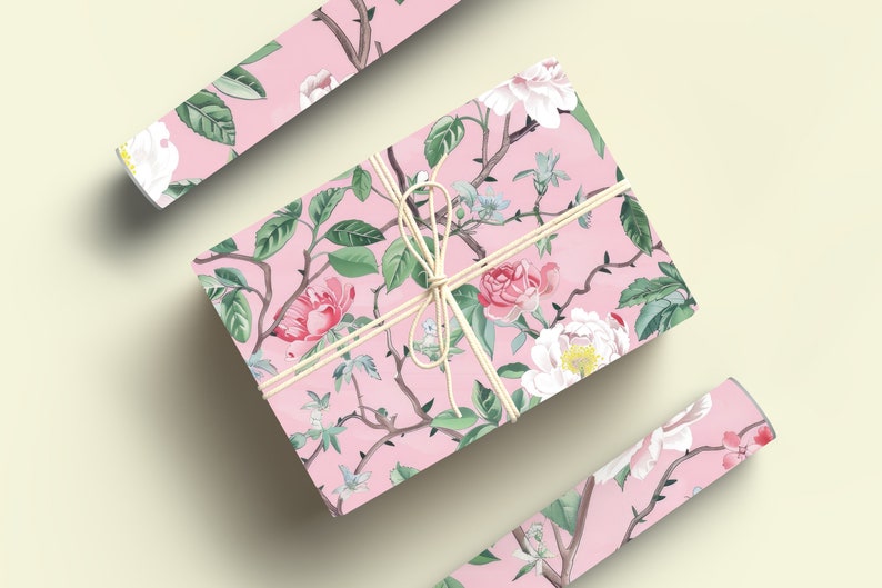 May include: A wrapped gift box and two rolls of wrapping paper with a pink floral pattern. The design features pink and white flowers, green leaves, and brown branches. The box is tied with twine.