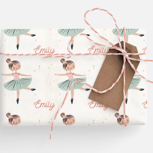 May include: White wrapping paper with a repeating pattern of a ballerina in a pink leotard and green tutu. The ballerina is holding her arms out to the side. The name "Emily" is written in pink cursive on the paper.