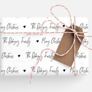 May include: White wrapping paper with a repeating pattern of black text that says "The Rodriguez Family" and "Merry Christmas" with a small black heart between each phrase. The paper is wrapped around a gift box with a brown gift tag tied with a pink and white twine.