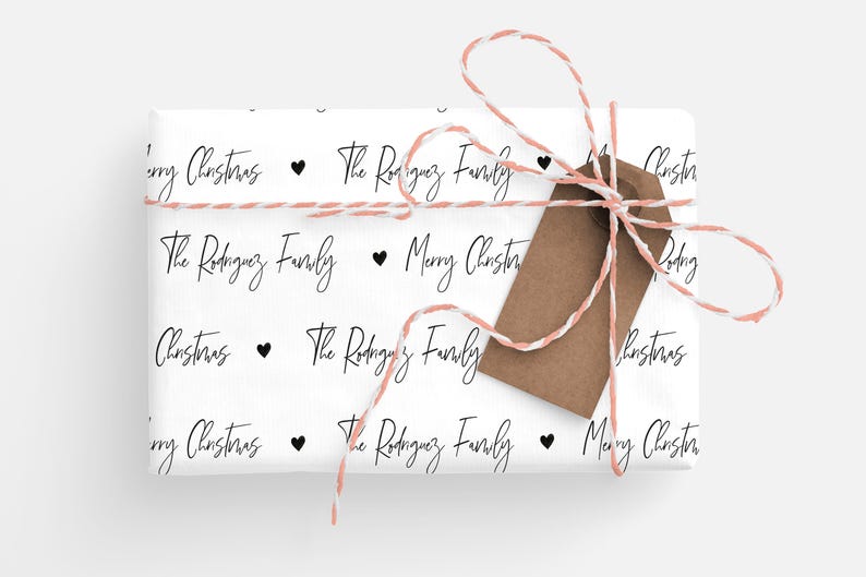 May include: White wrapping paper with a repeating pattern of black text that says "The Rodriguez Family" and "Merry Christmas" with a small black heart between each phrase. The paper is wrapped around a gift box with a brown gift tag tied with a pink and white twine.