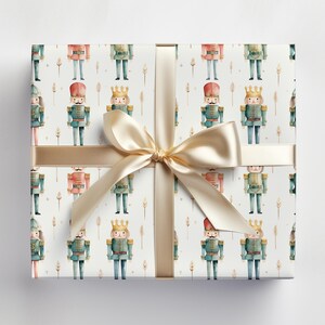 May include: White wrapping paper with a repeating pattern of watercolor nutcracker soldiers. The nutcrackers are wearing red, green, and blue uniforms with gold accents. A gold ribbon is tied around the paper.