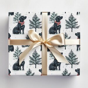 K&ouml;nnte beinhalten: Verpacktes Geschenk mit wei&szlig;em Hintergrund, das ein sich wiederholendes Muster aus schwarzen Labrador-Retrievern mit roten Schals und gr&uuml;nen Weihnachtsb&auml;umen zeigt. Ein goldenes Band und eine Schleife sind um das Geschenk gebunden.