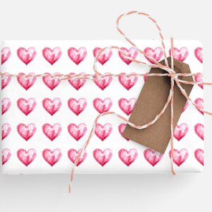 May include: A white gift box wrapped in pink watercolor heart patterned paper with a brown gift tag and tied with a pink and white twine bow.