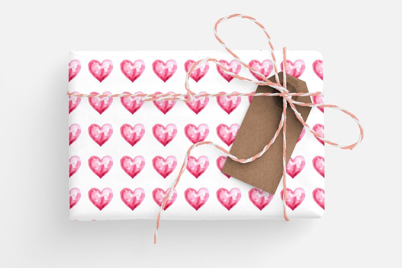 May include: A white gift box wrapped in pink watercolor heart patterned paper with a brown gift tag and tied with a pink and white twine bow.