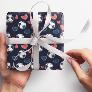 K&ouml;nnte beinhalten: Ein Geschenk, das in blaues Geschenkpapier mit einem wei&szlig;en Band eingewickelt ist. Das Papier hat ein Muster aus wei&szlig;en Fu&szlig;b&auml;llen und rosa Herzen.