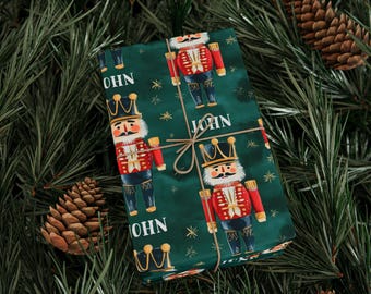 May include: A wrapped gift with a green background featuring a repeating pattern of nutcracker soldiers in red and blue uniforms, with the name "JOHN" printed on the wrapping paper. The gift is tied with twine and surrounded by pine needles and pine cones.