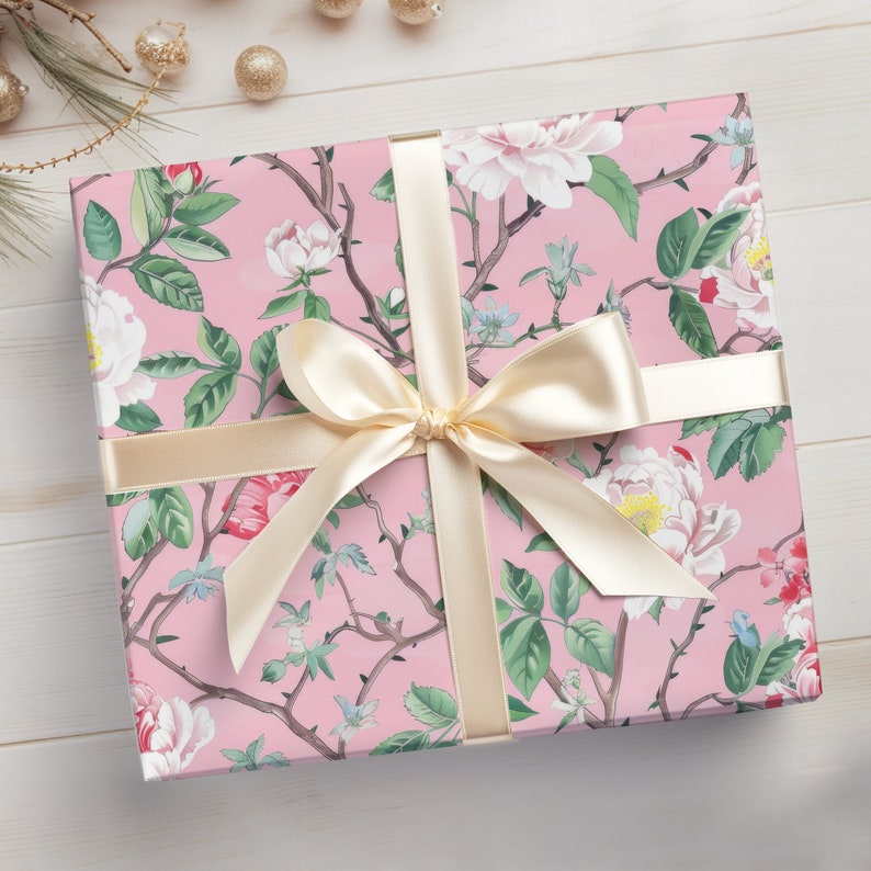 May include: A wrapped gift box featuring a pink floral pattern with white and red flowers, green leaves, and brown branches. The box is tied with a cream-colored ribbon and bow. The overall theme is gift-giving and celebration.