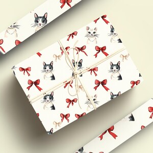 May include: A wrapped gift box and two rolls of wrapping paper with a repeating pattern of watercolour cat faces and red bows on a white background. The gift box is tied with twine.