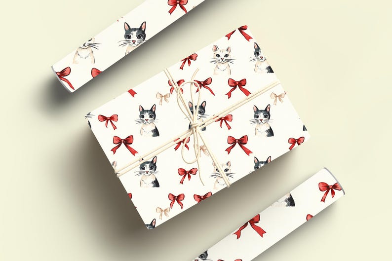 May include: A wrapped gift box and two rolls of wrapping paper with a repeating pattern of watercolour cat faces and red bows on a white background. The gift box is tied with twine.