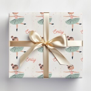 May include: White gift wrap with a repeating pattern of ballerinas in pink and green tutus. The name "Emily" is printed on the wrapping paper. A gold ribbon is tied around the box.