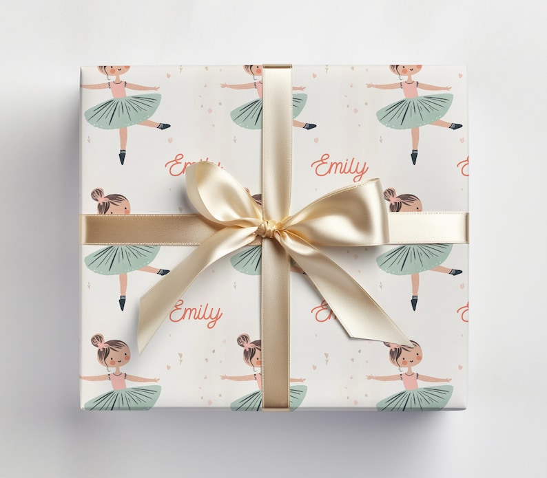 May include: White gift wrap with a repeating pattern of ballerinas in pink and green tutus. The name "Emily" is printed on the wrapping paper. A gold ribbon is tied around the box.