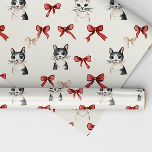 May include: Wrapping paper with a repeating pattern of watercolour cat portraits and red bows on a cream background. A roll of the paper is visible, partially unrolled, showing the design. The cats are in various poses.