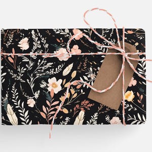Op de afbeelding: Een zwarte cadeaudoos met een bloemenpatroon in roze, wit en gele tinten. De doos is vastgebonden met een wit en roze touwtje en heeft een bruin papieren label.
