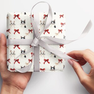May include: A wrapped gift with cream-coloured paper patterned with watercolour cats and red bows. It is tied with a grey satin ribbon. The hands are holding the gift.