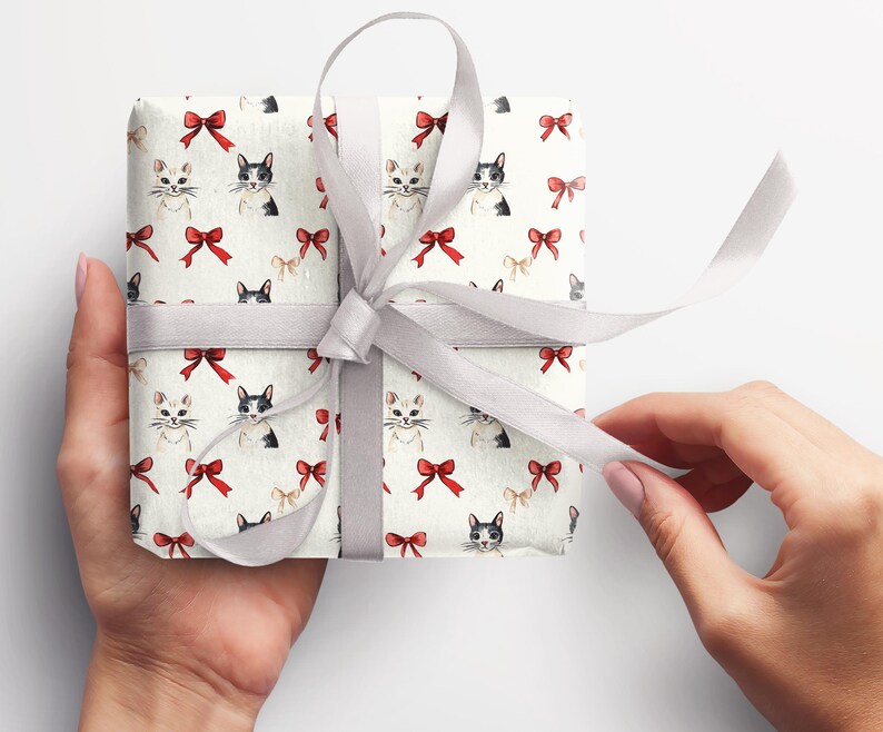 May include: A wrapped gift with cream-coloured paper patterned with watercolour cats and red bows. It is tied with a grey satin ribbon. The hands are holding the gift.