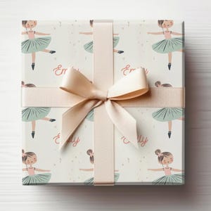 May include: A white gift box with a pink and green ballerina pattern. The box is wrapped with a beige ribbon and bow. The text "Emily" is repeated on the box.