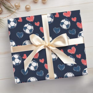 K&ouml;nnte beinhalten: Blaue Geschenkpapier mit einem wei&szlig;en Band und einem Muster aus roten Herzen und wei&szlig;en Fu&szlig;b&auml;llen.