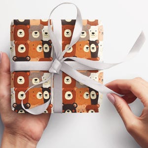Op de afbeelding: Wit cadeaupapier met een herhalend patroon van bruine, zwarte en witte beren. De beren kijken allemaal naar voren en hebben verschillende gezichtsuitdrukkingen. Het cadeaupapier is vastgebonden met een wit lint.