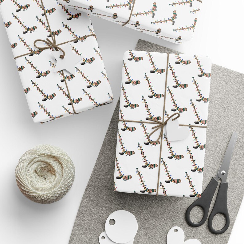 K&ouml;nnte beinhalten: Wei&szlig;es Geschenkpapier mit einem sich wiederholenden Muster aus Hockeyschl&auml;gern, die mit bunten Weihnachtslichtern verziert sind. Das Papier ist um eine Geschenkbox mit einer braunen Schnur gebunden.