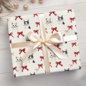 May include: A wrapped gift with cream-coloured paper decorated with cat faces and red bows. A cream-coloured ribbon and bow are tied around the package. The gift is on a white wooden surface.
