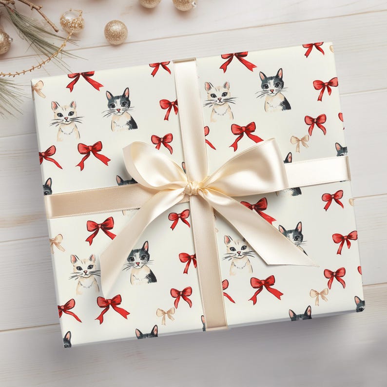 May include: A wrapped gift with cream-coloured paper decorated with cat faces and red bows. A cream-coloured ribbon and bow are tied around the package. The gift is on a white wooden surface.