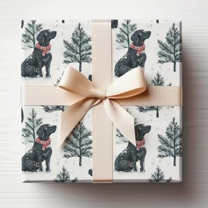 K&ouml;nnte beinhalten: Eine Geschenkbox mit einem sich wiederholenden Muster aus schwarzen Labrador-Hunden mit roten Schottenkaro-Schals, die mit stilisierten immergr&uuml;nen B&auml;umen auf wei&szlig;em Hintergrund durchsetzt sind. Die Box ist mit einem beigen Band und einer Schleife gebunden.