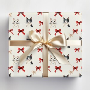 May include: A cream-coloured gift box featuring a pattern of cats and bows. The box is wrapped with a gold ribbon and bow. The pattern includes watercolour illustrations of cats in black and white, and red bows.