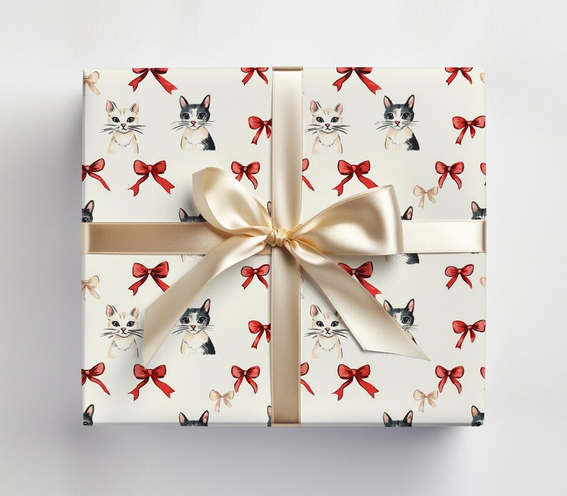 May include: A cream-coloured gift box featuring a pattern of cats and bows. The box is wrapped with a gold ribbon and bow. The pattern includes watercolour illustrations of cats in black and white, and red bows.