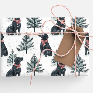 K&ouml;nnte beinhalten: Geschenkpapier mit einem sich wiederholenden Muster aus schwarzen Hunden mit roten Schottenkaro-Schals und Tannen auf wei&szlig;em Hintergrund. Gebunden mit rosa-wei&szlig;em Garn und einem braunen Anh&auml;nger. Festliches Design.