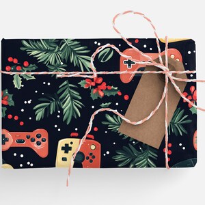 K&ouml;nnte beinhalten: Ein Geschenk mit festlichem Design. Das Geschenkpapier zeigt ein Muster aus roten und gelben Gamecontrollern, gr&uuml;nen Tannenzweigen, roten Beeren und wei&szlig;en Punkten auf einem dunkelblauen Hintergrund. Ein braunes Etikett und rosa-wei&szlig;es Garn vervollst&auml;ndigen das Geschenk.