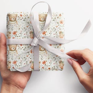 May include: A white gift box with a silver ribbon. The box is wrapped in paper with a repeating pattern of brown bears wearing sweaters and flowers.