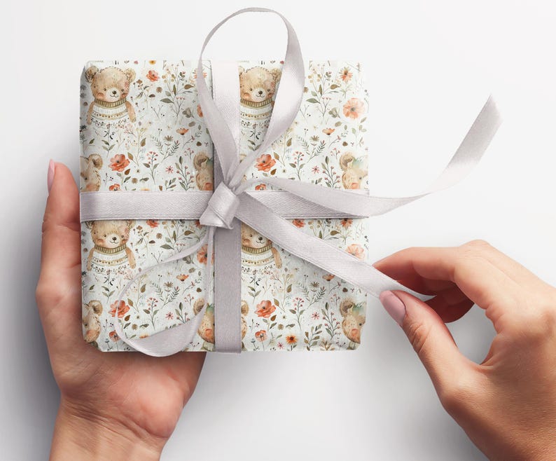May include: A white gift box with a silver ribbon. The box is wrapped in paper with a repeating pattern of brown bears wearing sweaters and flowers.