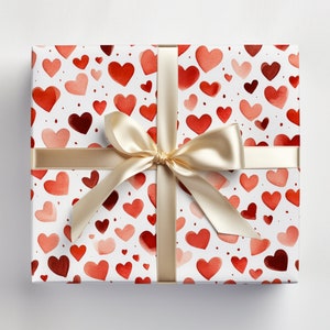 Op de afbeelding: Een witte cadeaudoos met een rood en roze aquarelhartpatroon. De doos is vastgebonden met een cr&egrave;mekleurig satijnen lint met een strik.