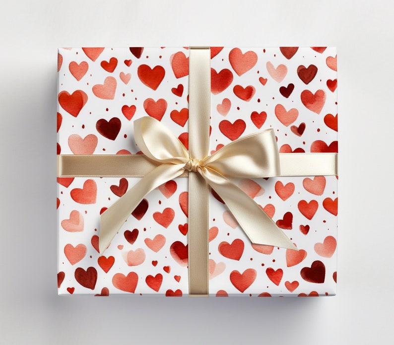 Op de afbeelding: Een witte cadeaudoos met een rood en roze aquarelhartpatroon. De doos is vastgebonden met een cr&egrave;mekleurig satijnen lint met een strik.