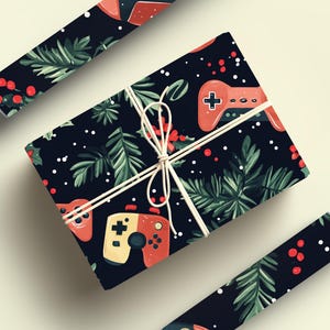 K&ouml;nnte beinhalten: Geschenk verpackt in schwarzem Papier mit einem Muster aus roten Gamecontrollern, gr&uuml;nen Tannenzweigen und roten Beeren. Das Geschenk ist mit wei&szlig;em Bindfaden gebunden. Zwei Rollen passendes Geschenkpapier sind ebenfalls sichtbar.