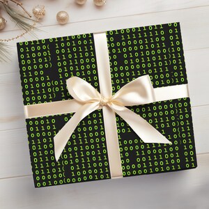Peut inclure: Un cadeau emball&eacute; avec du papier noir imprim&eacute; d'un motif de code binaire vert. Il est nou&eacute; avec un ruban et un n&oelig;ud en satin cr&egrave;me. Le cadeau est pos&eacute; sur une surface en bois blanc.