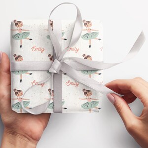 May include: White gift wrap with a repeating pattern of a ballerina in a green tutu. The name "Emily" is printed in pink on the wrapping paper.